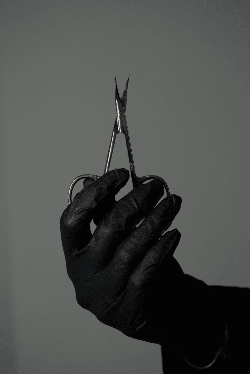 A black gloved hand holds small scissors against a gray background.