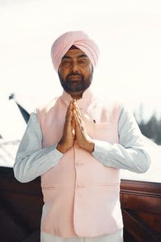 A man wearing a pink turban and vest stands outdoors with praying hands. PNG