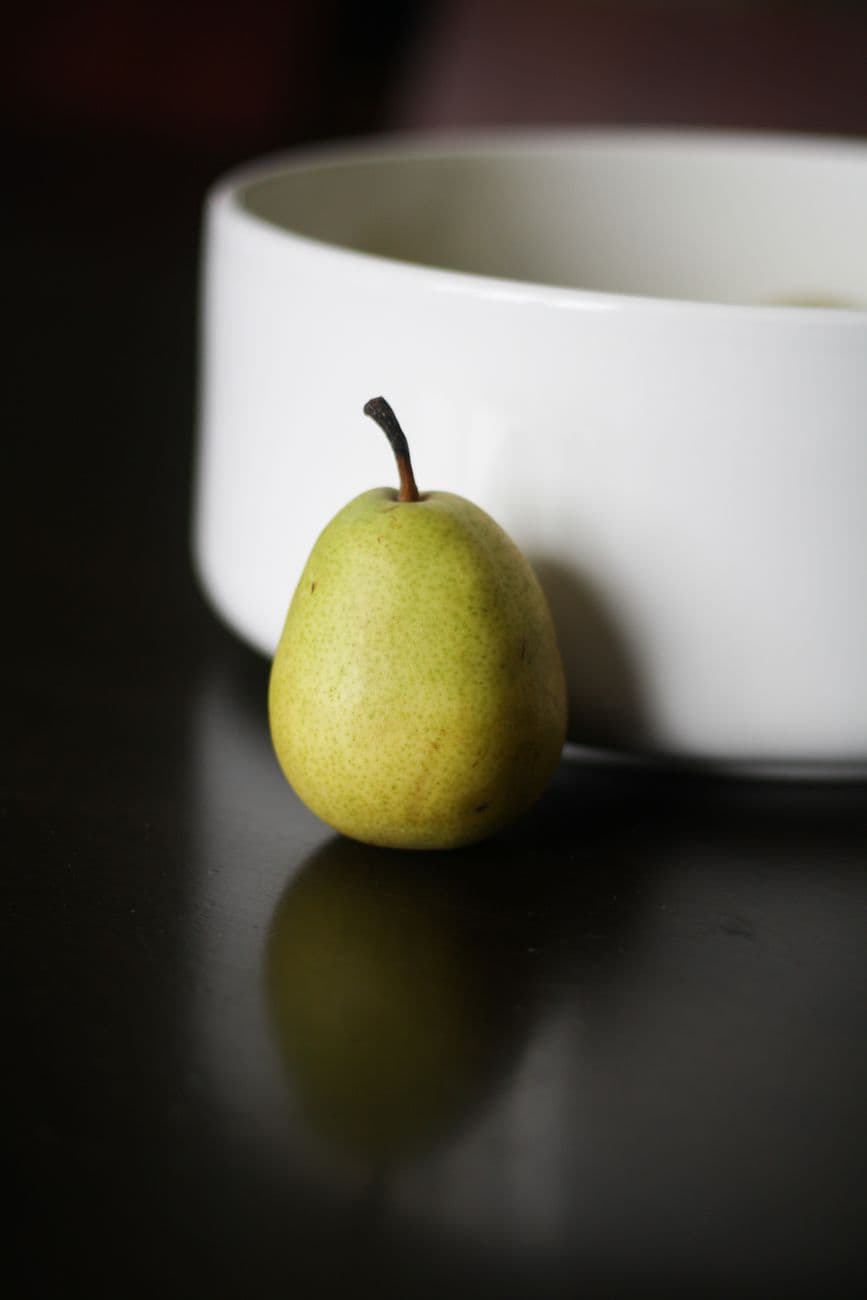 A ripe green pear placed beside a white bowl on a dark reflective surface.