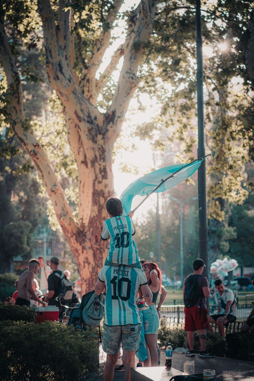 Father and son in Argentina soccer jerseys celebrate outdoors with a flag.