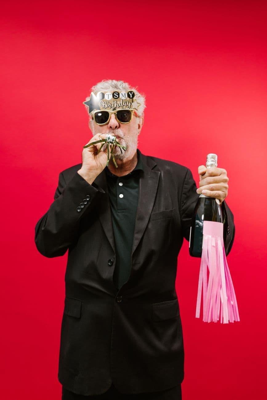 Senior man with party props celebrating birthday against red background.