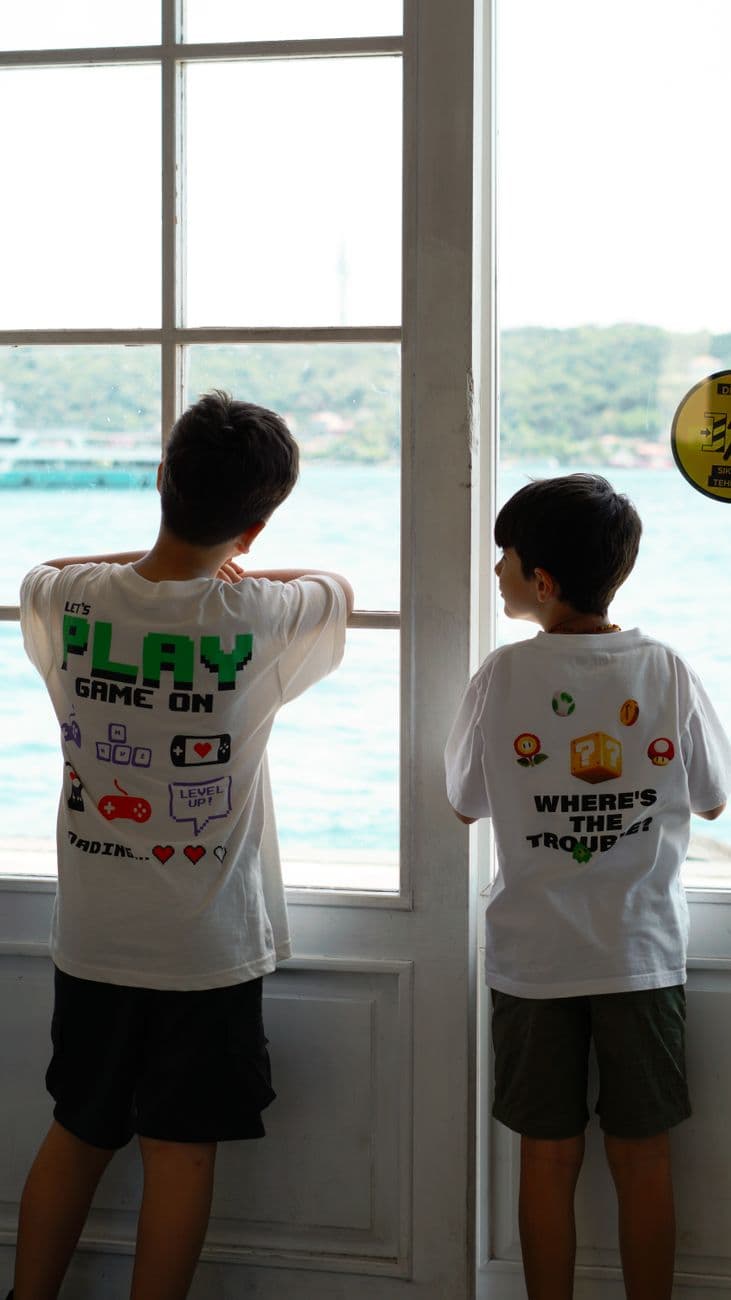 Two children with graphic T-shirts stand by a window, peering out at the sea.