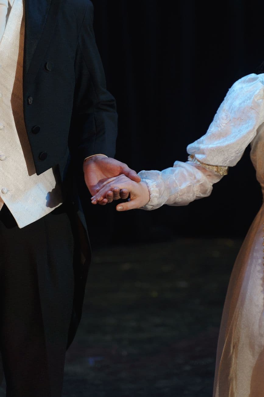 A close-up of two costumed performers holding hands on stage, evoking romance in a theatri