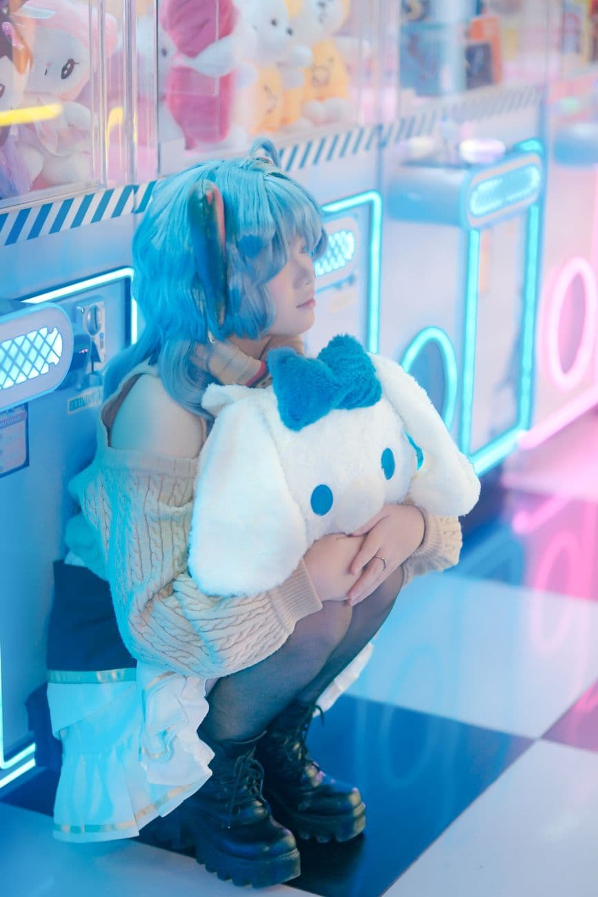 A cosplayer with blue hair sits holding a plush toy in a neon-lit arcade environment.