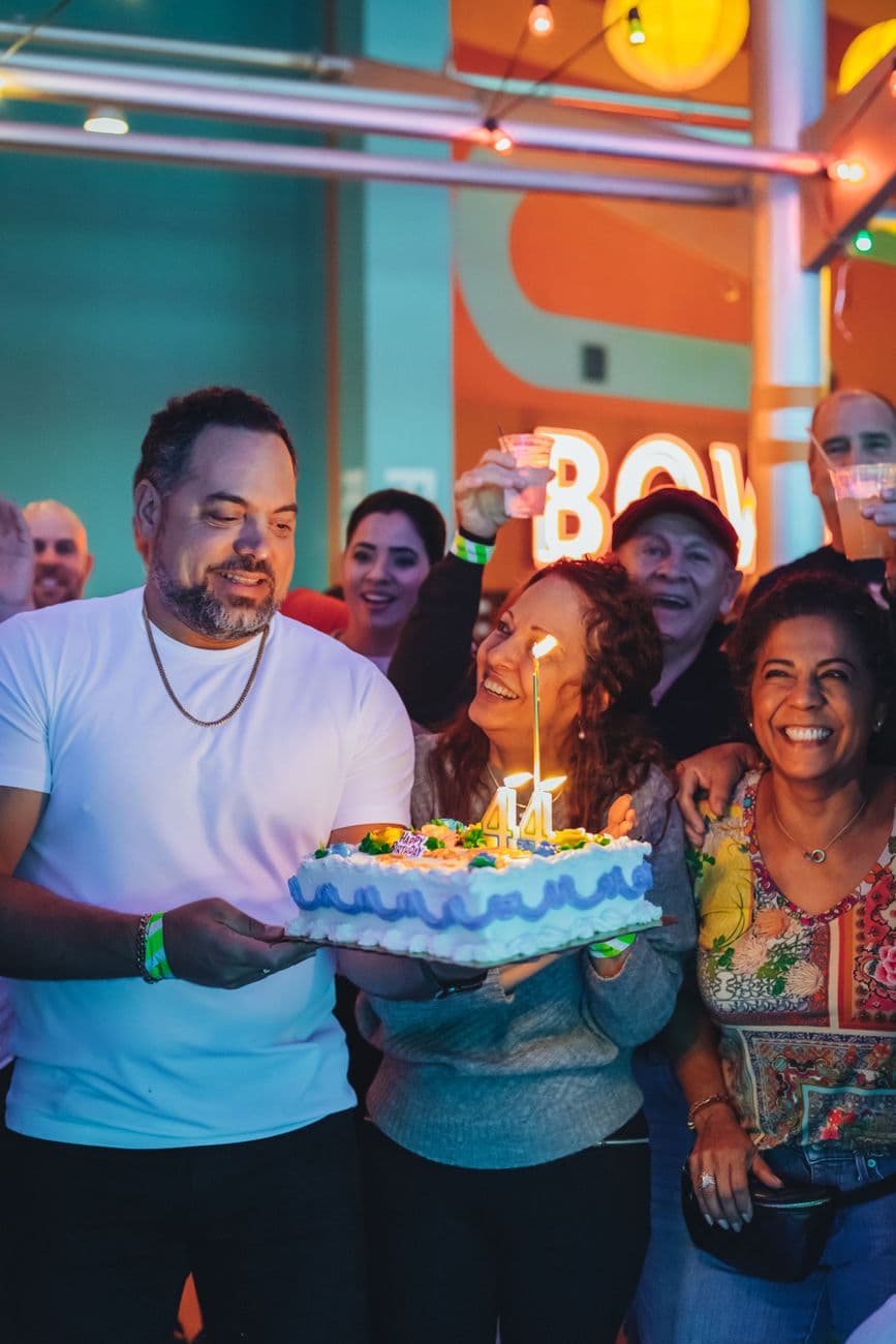A lively birthday celebration with smiling friends and a candle-lit cake indoors.