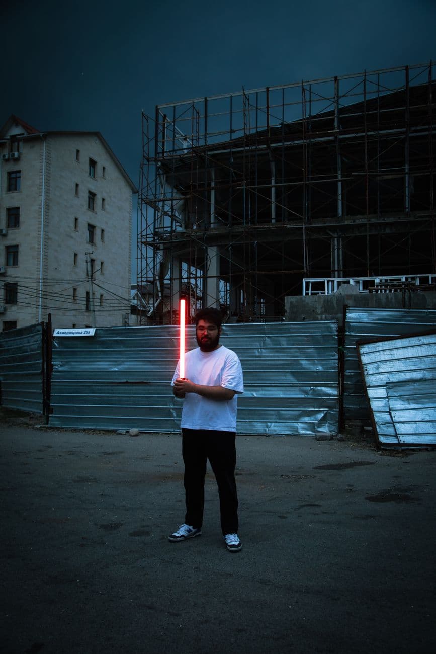 A man holding a neon light stands in front of a construction site at dusk, creating a mood