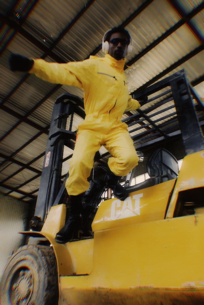 A man in a vibrant yellow jumpsuit poses energetically on a forklift inside a warehouse.