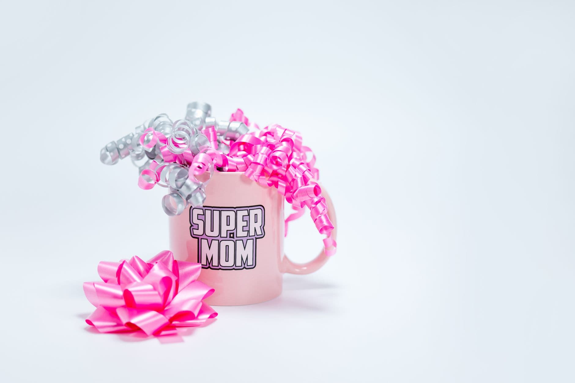 A pink Super Mom mug adorned with curly pink and silver ribbons on a white background, per