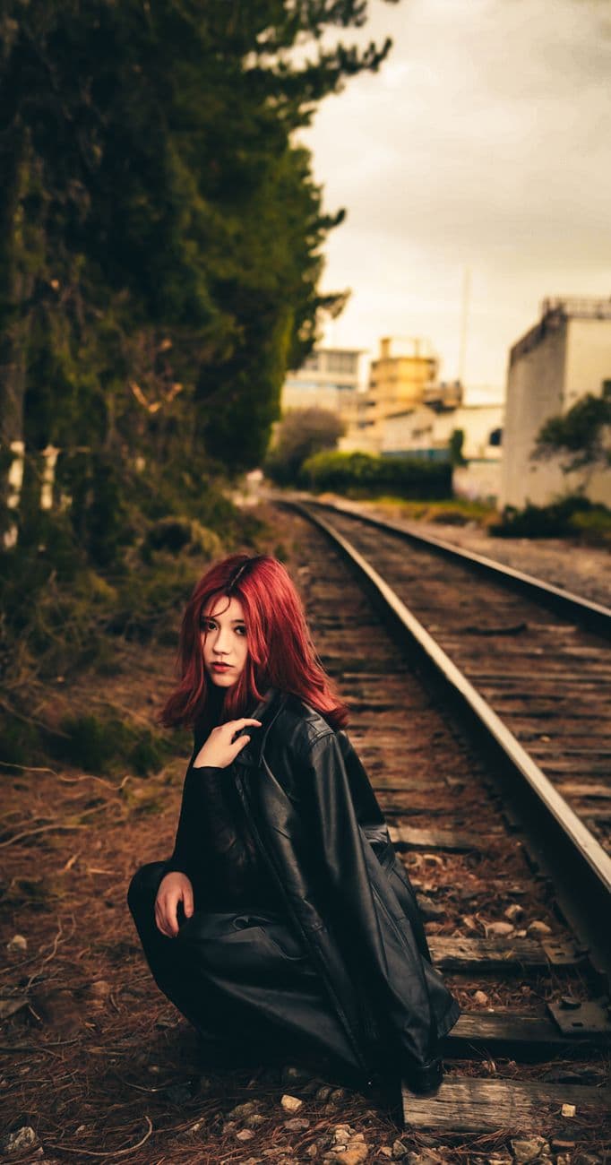 A red-haired woman in a black jacket crouches beside railway tracks amidst urban greenery.