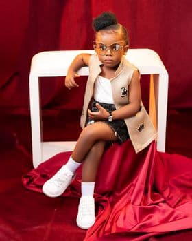 A stylish young girl poses confidently in a chic outfit with glasses and crossed legs. PNG