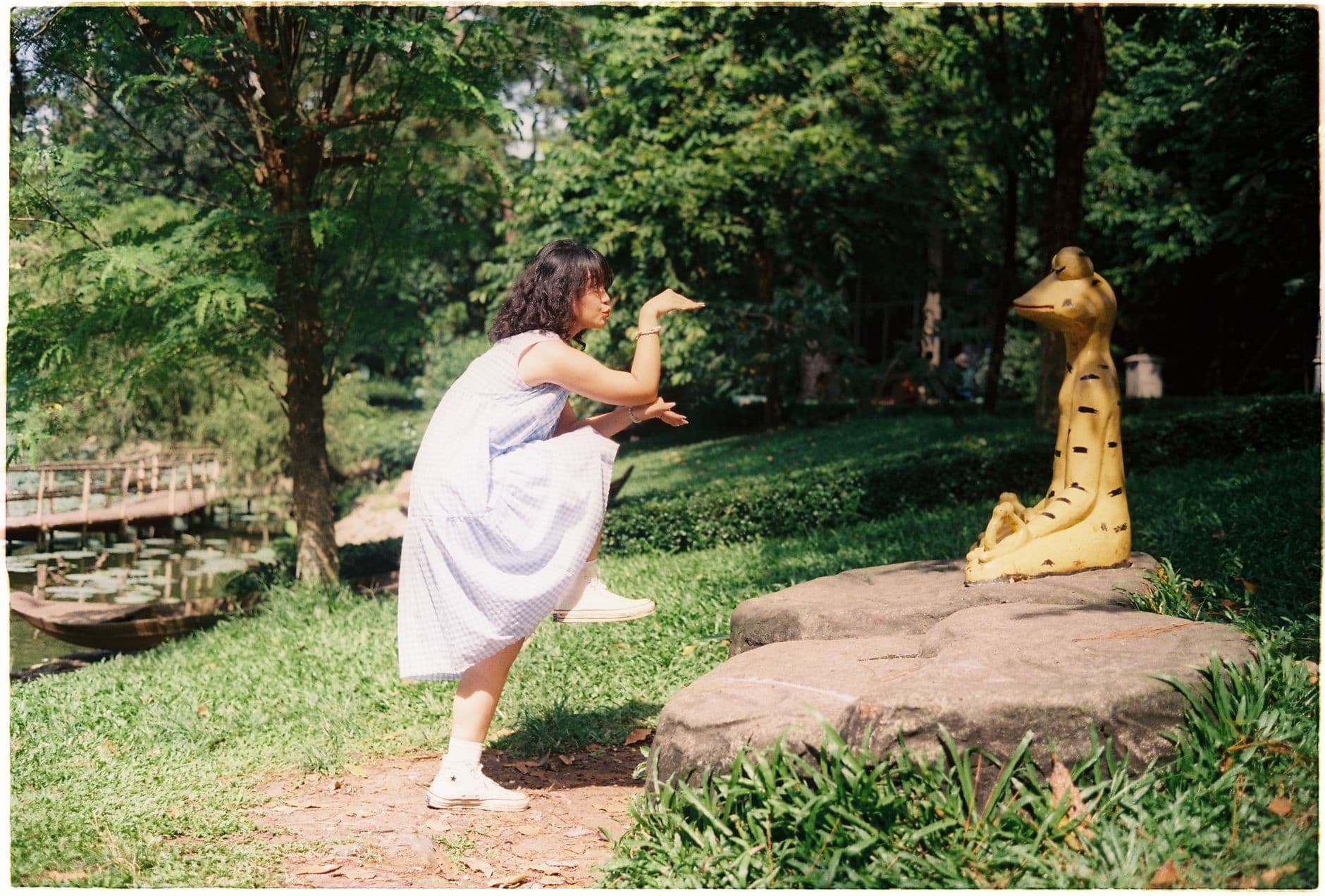 A woman playfully mimics a snake statue in a sunlit park, surrounded by greenery.