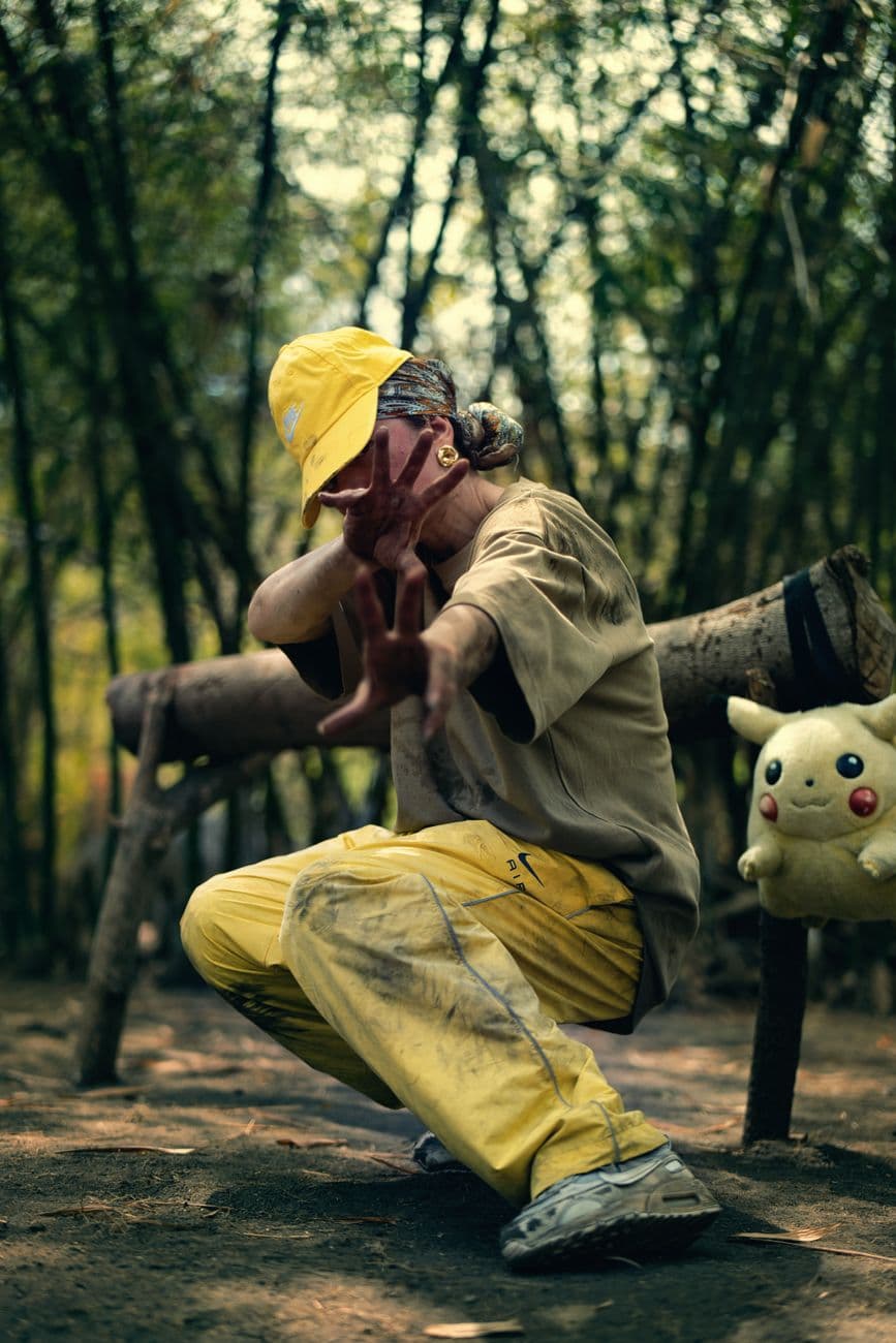 A young adult wears casual attire, poses outdoors in a bamboo forest beside a plush toy, e