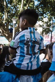 A young boy wearing a Messi jersey sits on shoulders in a festive crowd in Buenos Aires. PNG
