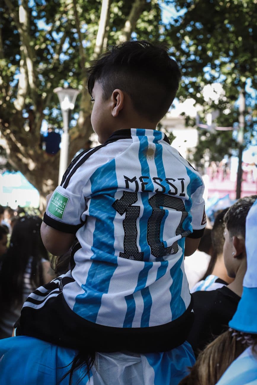 A young boy wearing a Messi jersey sits on shoulders in a festive crowd in Buenos Aires.