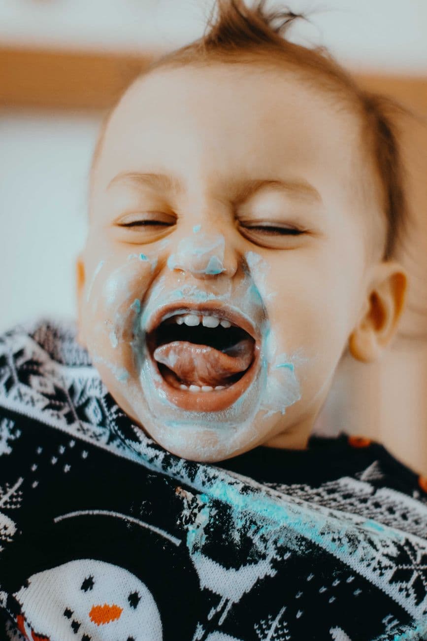 Adorable toddler laughing with icing on face, wearing festive Christmas sweater indoors. C