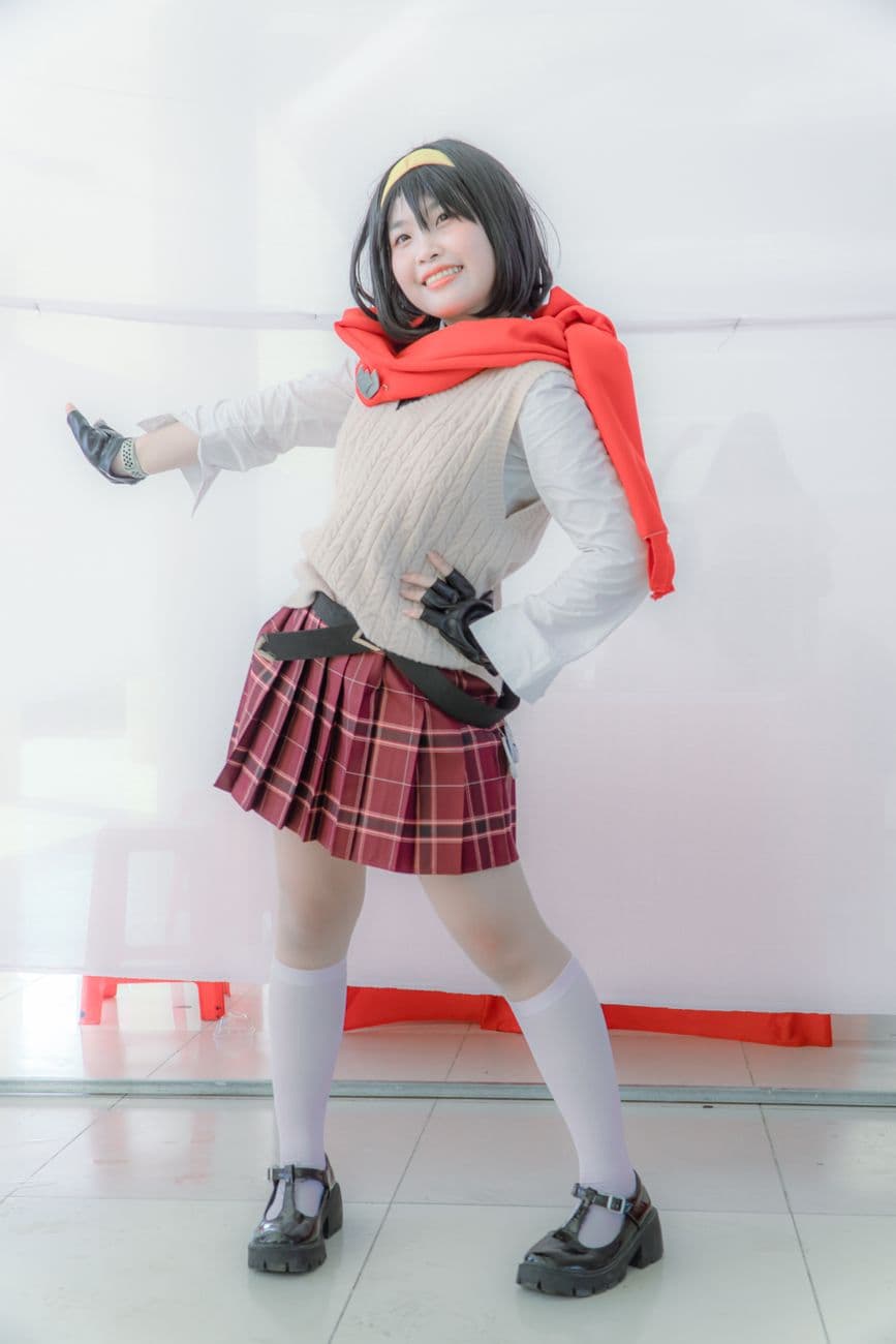 Cheerful cosplayer posing in a colorful anime costume with plaid skirt and red scarf.