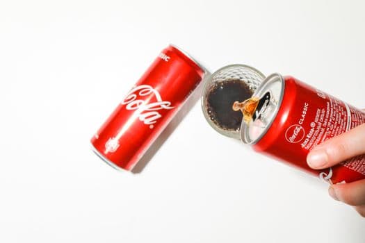 Close-up of cola being poured from a red can into a glass against a white background. PNG