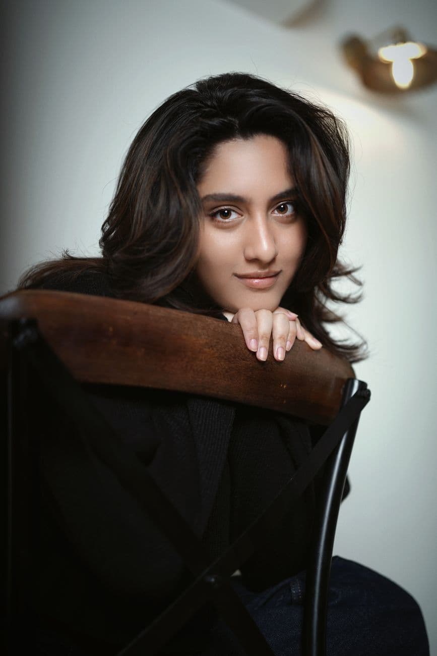 Elegant portrait of a young woman leaning on a wooden chair indoors with soft lighting.