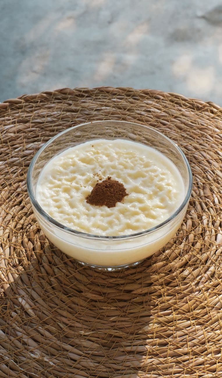 Glass bowl of Turkish sütlaç dessert with cinnamon, set on woven mat, natural lighting.