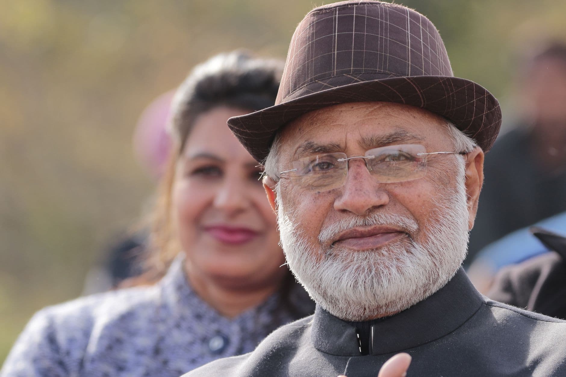 Portrait of an elderly man with a white beard wearing a hat outdoors with a woman in the b