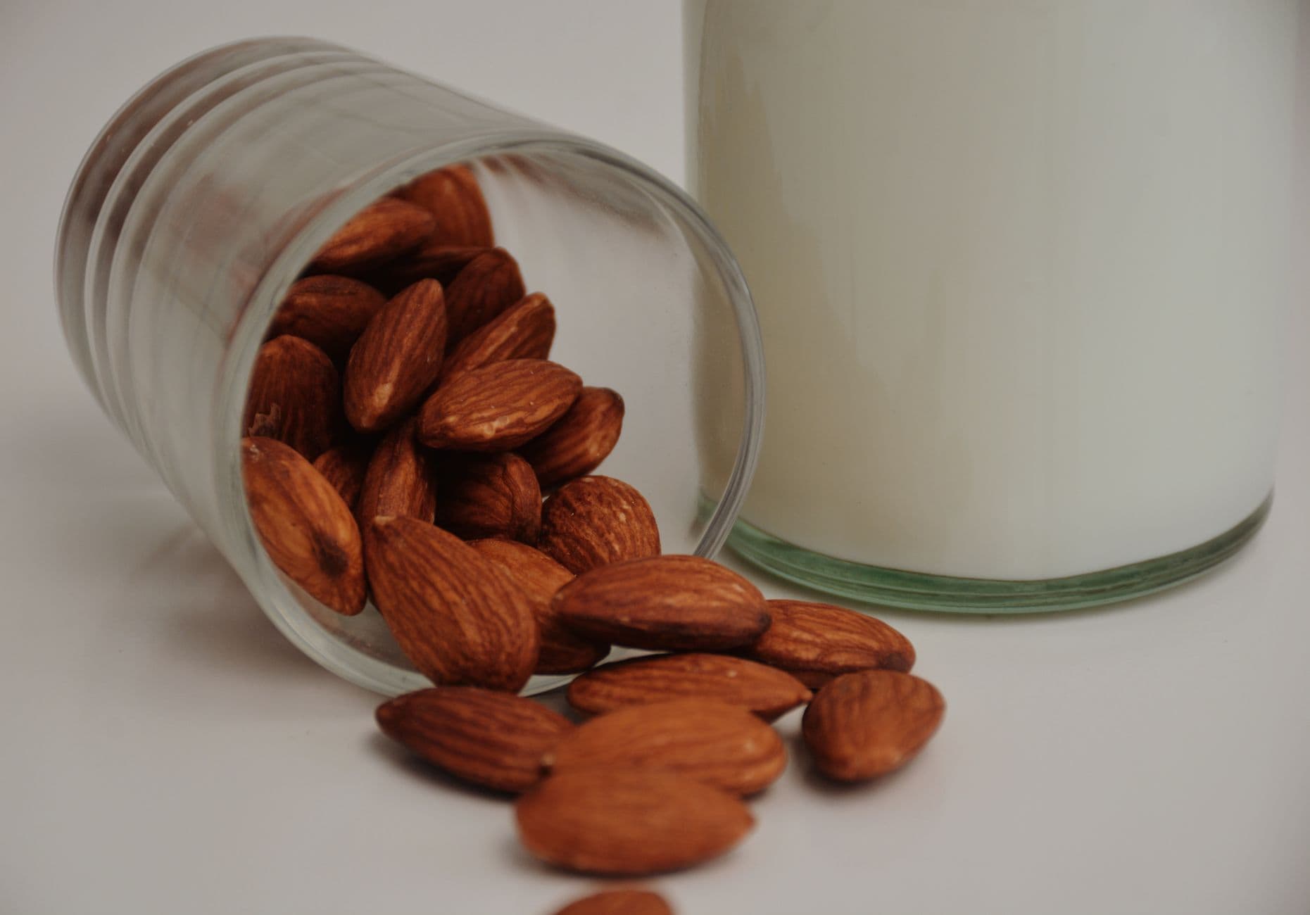 Transparent glass with spilled whole crispy almonds near dairy beverage on white backgroun