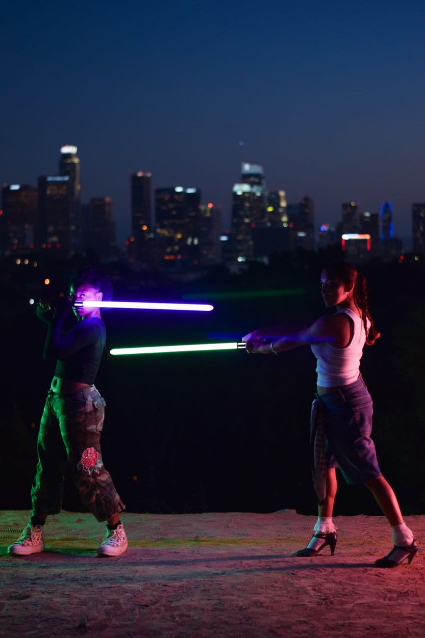 Two people engage in a lightsaber duel with the Los Angeles cityscape at dusk.