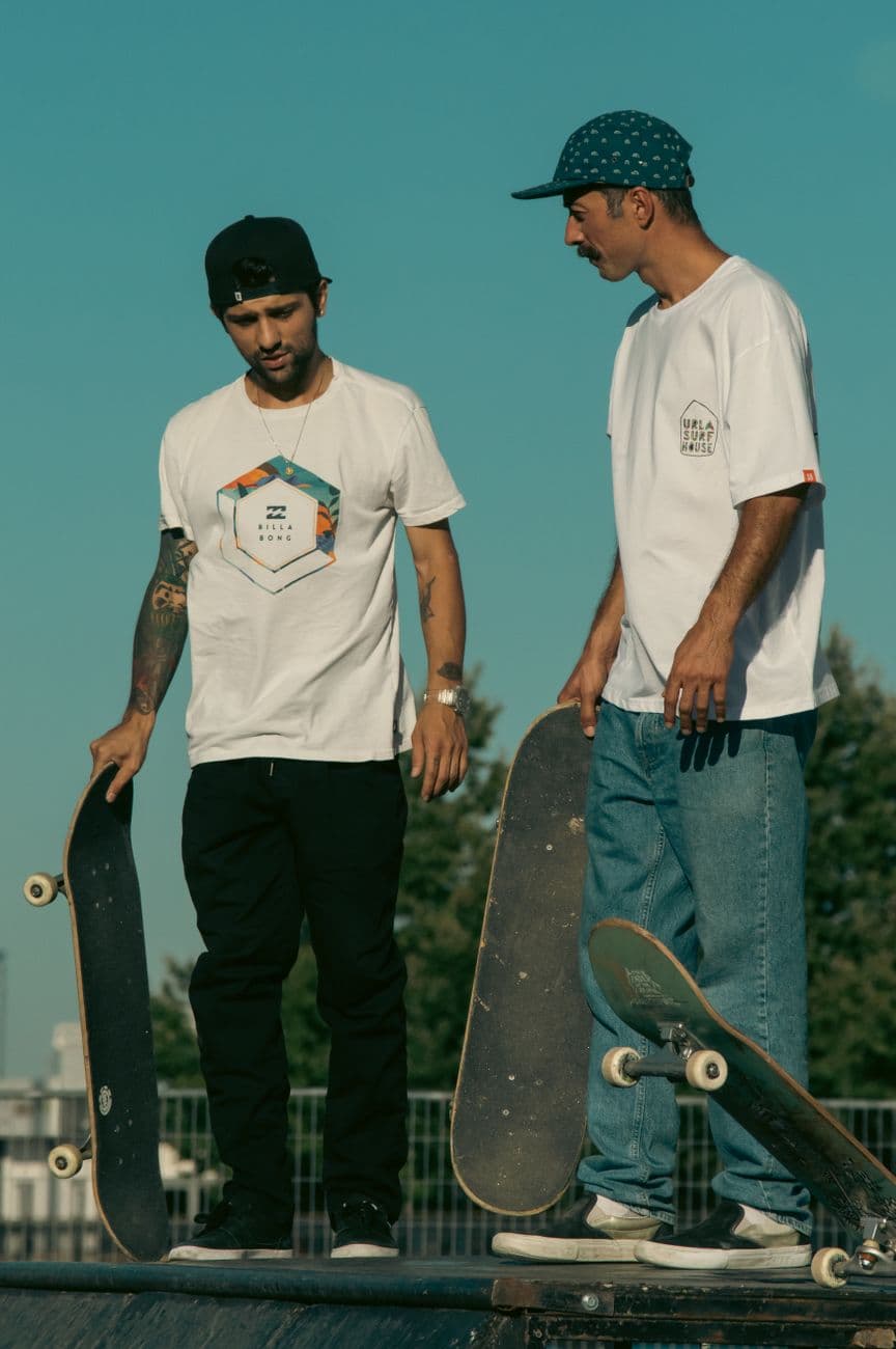 Two skateboarders in casual clothing chat at a skatepark, holding skateboards.