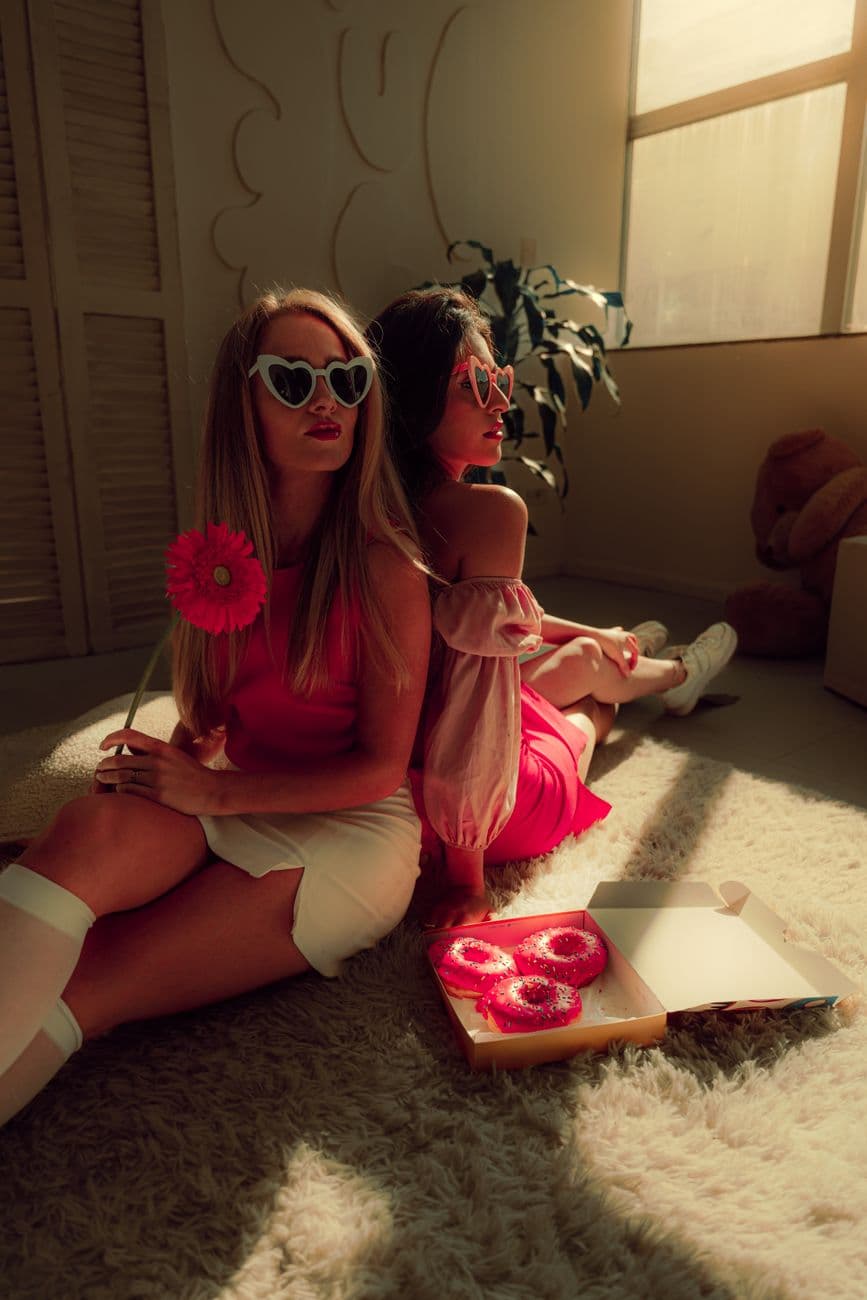 Two stylish women with heart-shaped sunglasses enjoy pink donuts in a cozy sunlit room.